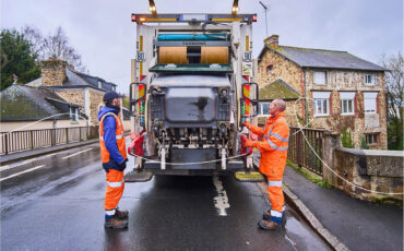 2 rippeurs sont à l'arrière d'une benne à ordures. Un bac est en train de se vider dans celle-ci.