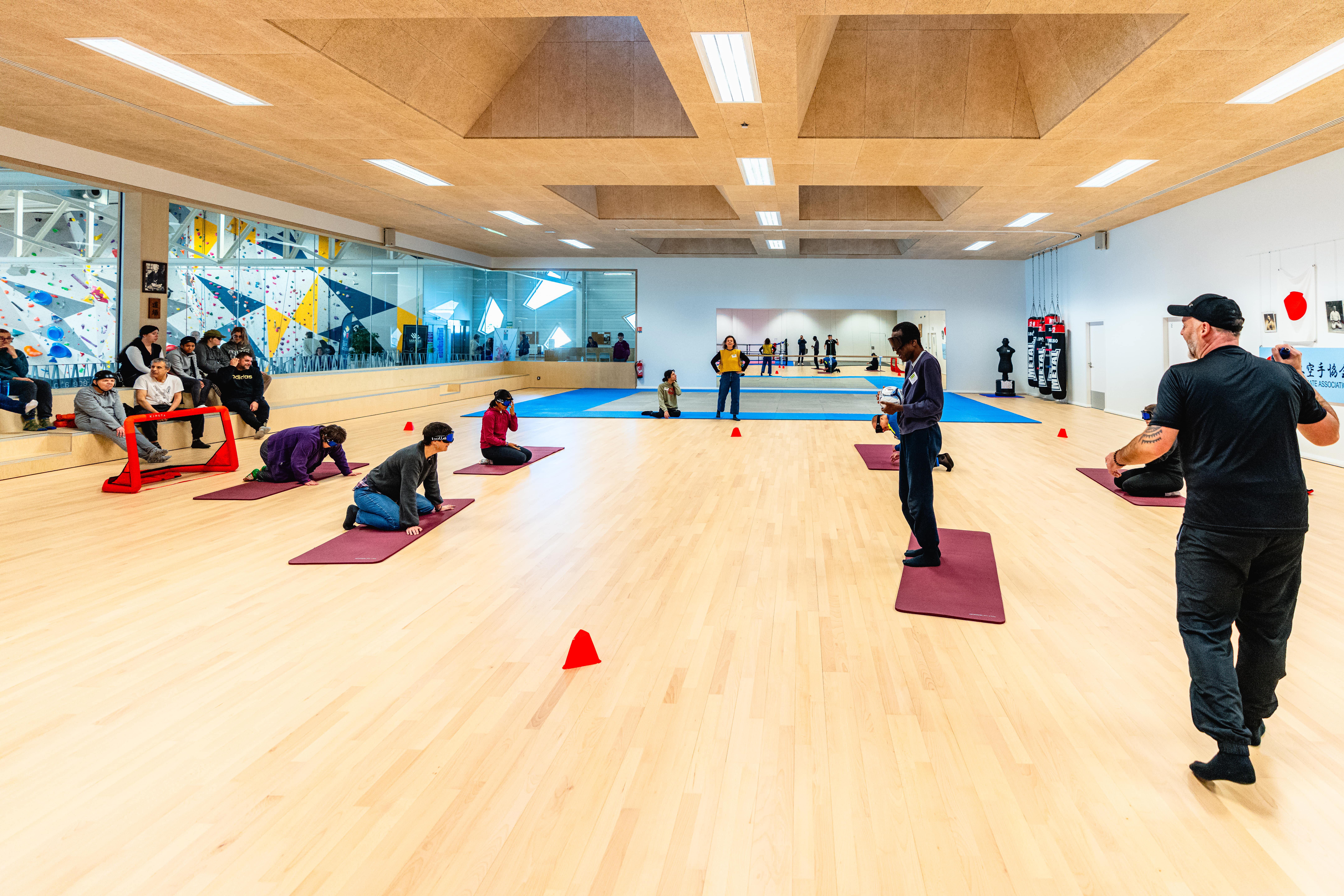 deux équipes de trois personnes jouent au torball. Une seule est debout et tiens la balle. Ils ont tous les yeux bandés.