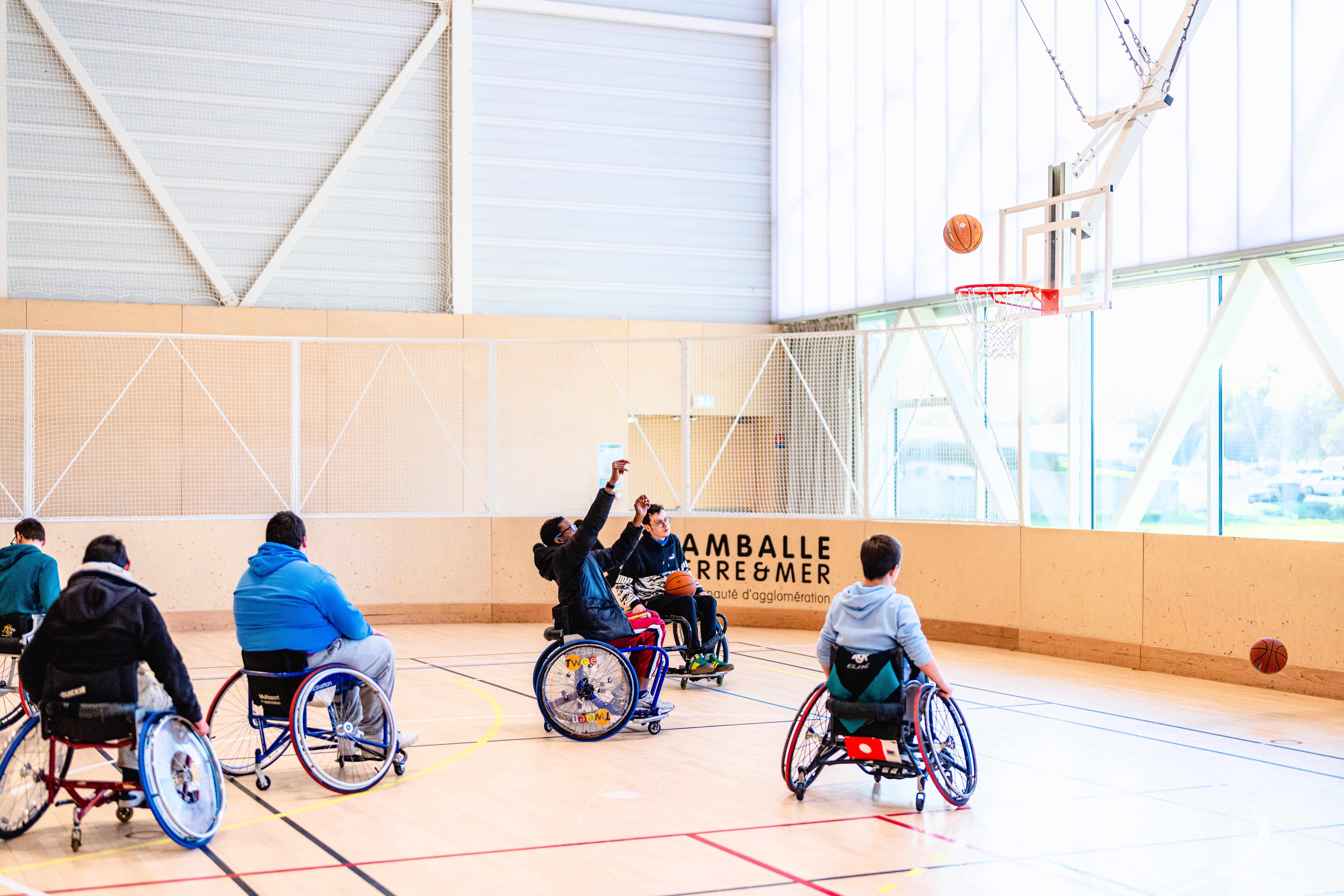 six personnes jouent au basket fauteuil. Le ballon est en l'air, prêt a arriver dans le panier.