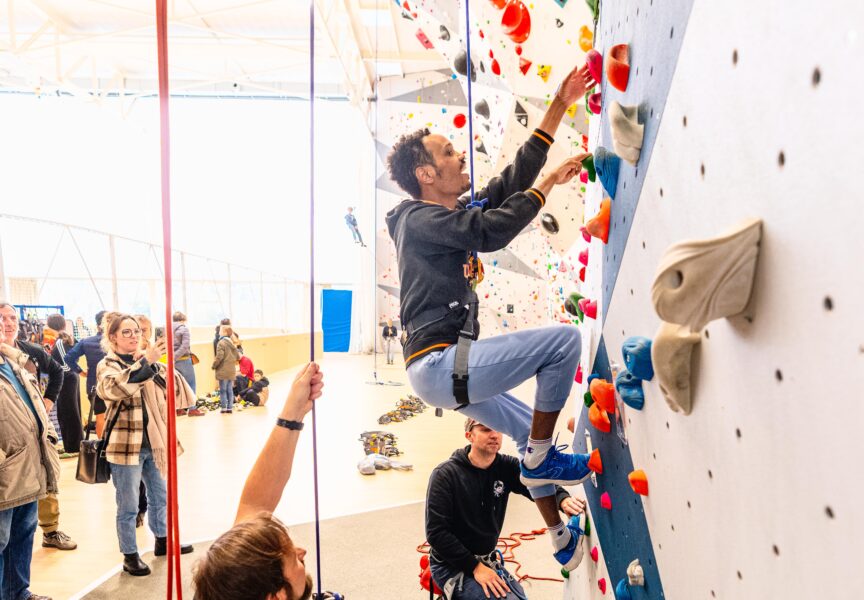une personne en situation de handicap grimpe le mur d'escalade et est assuré par un homme en bas.
